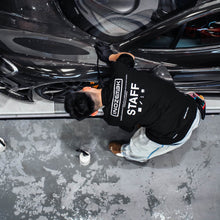 Person in a black 'Staff' sweatshirt working on a black car in a garage.